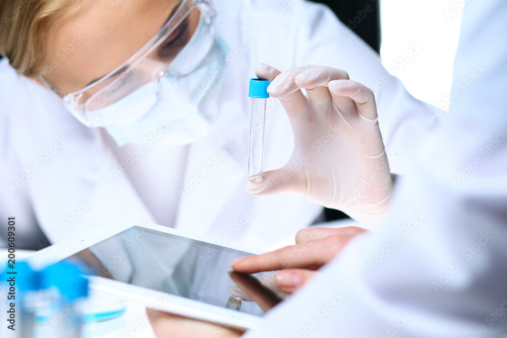 Closeup of scientific research team with clear solution in laboratory. Blonde female chemist holds test tube of glass while her colleague checks results with tablet pc. Blood test, medicine or Closeup of scientific research team with clear solution in laboratory. Blonde female chemist holds test tube of glass while her colleague checks results with tablet pc. Blood test, medicine or