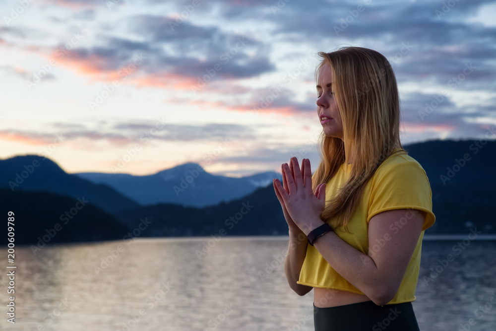 Woman in prayer position is meditating with her eyes closed during a vibrant sunset. Taken in Whytecliff Park, Horseshoe Bay, West Vancouver, British Columbia, Canada. Woman in prayer position is meditating with her eyes closed during a vibrant sunset. Taken in Whytecliff Park, Horseshoe Bay, West Vancouver, British Columbia, Canada.