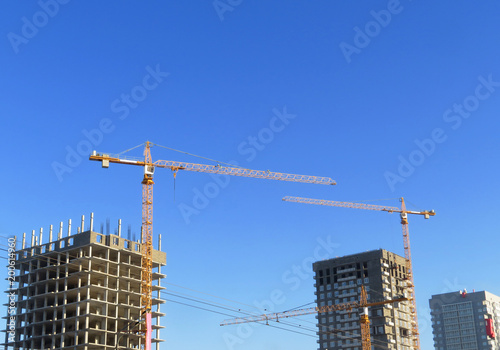 Three cranes building houses. Against a pure blue sky. Built a house and two unfinished high-rise buildings.