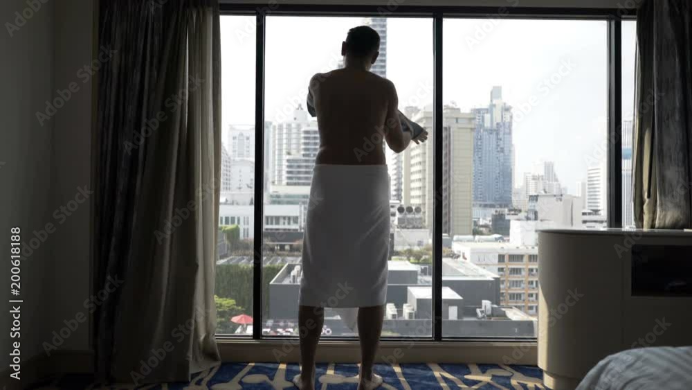 Man wearing towel and puts his shirt on while enjoying the view from the window
Man wearing towel and puts his shirt on while enjoying the view from the window