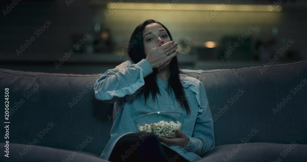 Sad, bored and depressed woman sitting on a sofa at home watching a bad movie eating popcorn