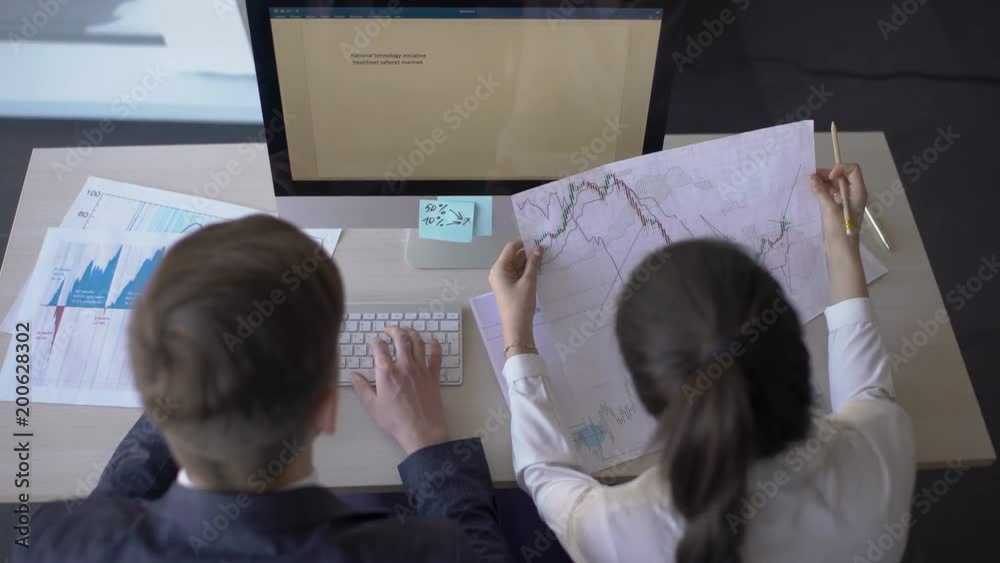 Two young business people are working on blueprint, sitting at table with PC, looking, reading documents, sharing ideas, talking at desk in large company. 4k Colleagues in formal attire analyze papers Two young business people are working on blueprint, sitting at table with PC, looking, reading documents, sharing ideas, talking at desk in large company. 4k Colleagues in formal attire analyze papers