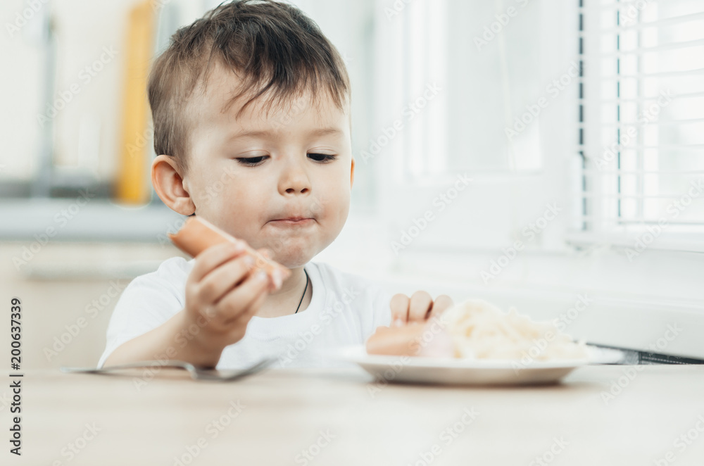 The child is sitting in the kitchen and greedily eats sausage The child is sitting in the kitchen and greedily eats sausage