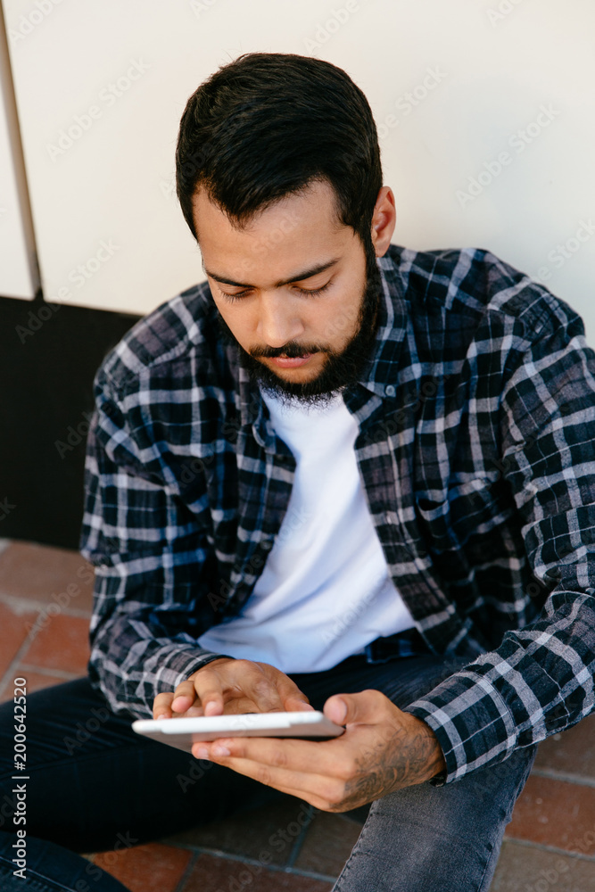 Pensive attractive man using a computer tablet, browsing websites, while sitting on the ground near the urban wall. Dressed up in stylish plaid shirt and jeans.
