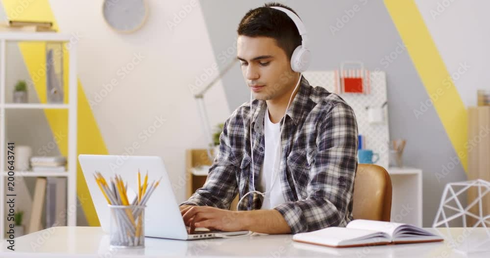 Caucasian young handsome man in the motley shirt and headphones taping and typing on the laptop computer while sitting at the desk in the modern office. Inside Caucasian young handsome man in the motley shirt and headphones taping and typing on the laptop computer while sitting at the desk in the modern office. Inside