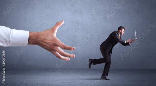 Young miniature businessman running from a big hand with blueish grey background