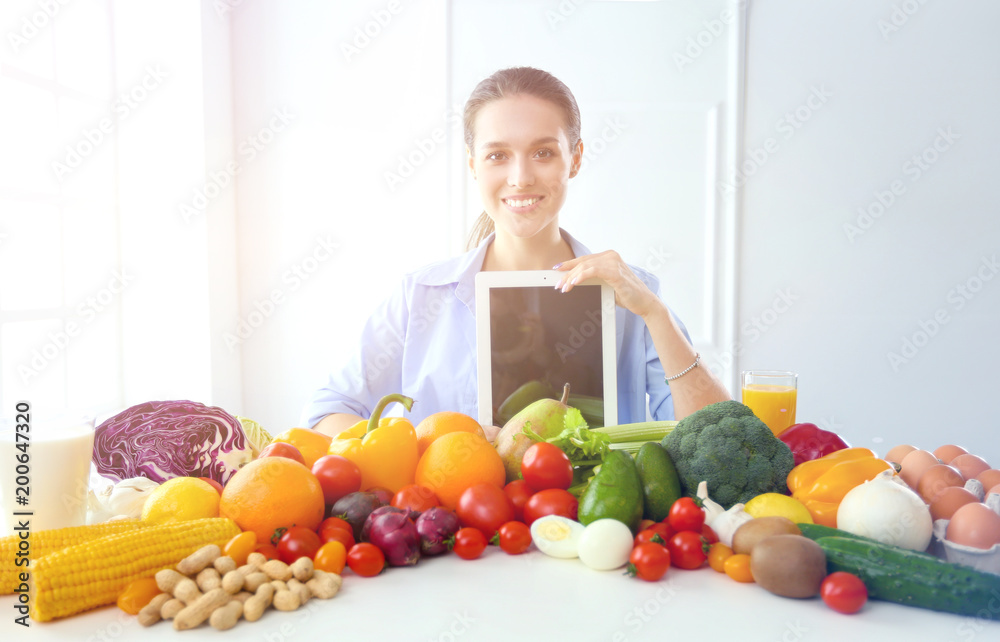 Young and cute woman sitting at the table full of fruits and vegetables in the wooden interior, holding tablet Young and cute woman sitting at the table full of fruits and vegetables in the wooden interior, holding tablet