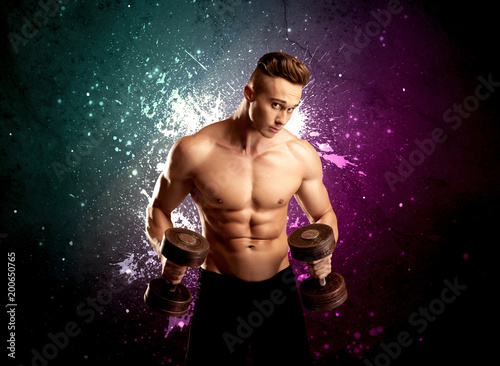 A sexy male fitness trainer showing his muscles and looking seductive with a weight in his hands in front of bright paint splash purple wall concept. © ra2 studio