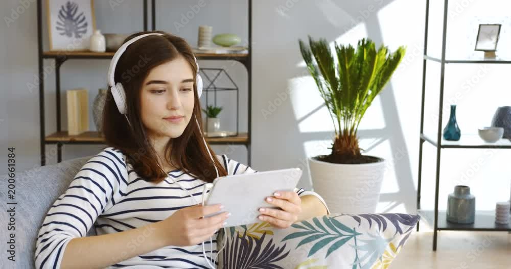 Young attractive woman in the striped blouse and big white headphones watching something on the tablet computer on the gray couch in the living room and smiling. Inside Young attractive woman in the striped blouse and big white headphones watching something on the tablet computer on the gray couch in the living room and smiling. Inside