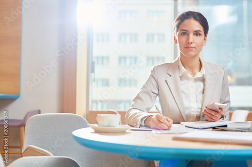 Young serious businesswoman looking at camera while sitting by table in cafe and making working notes