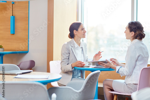 Two confident businesswomen sitting opposite one another by table in cafe and discussing working information for project