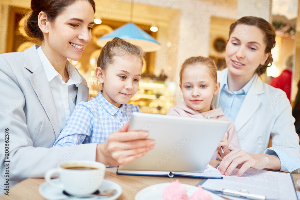 Fototapeta premium Two female employees and their daughters watching curious video in the net while spending time in cafe
