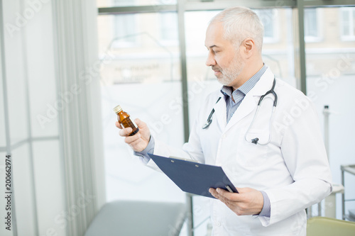 Grey-haired doctor looking at medicine in bottle in his hand while working in clinics