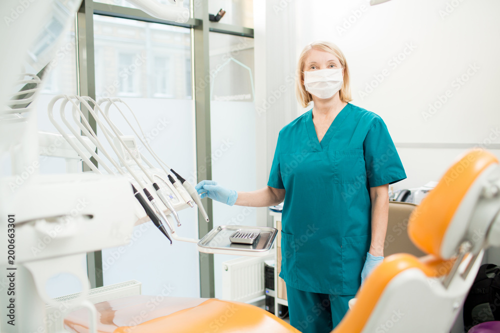 Nurse in uniform, mask and gloves looking at camera while standing by workplace of dentist Nurse in uniform, mask and gloves looking at camera while standing by workplace of dentist