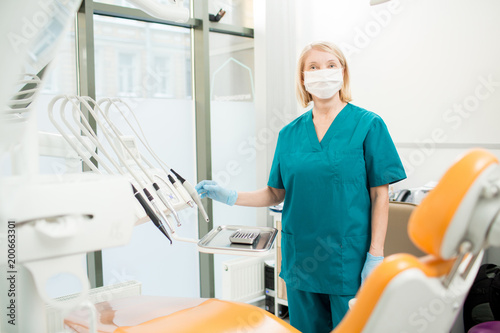 Nurse in uniform, mask and gloves looking at camera while standing by workplace of dentist