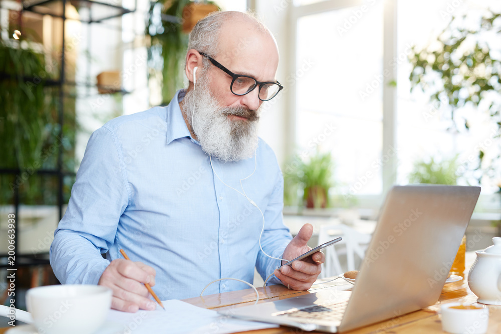 Serious mature bearded man with smartphone and earphones looking at laptop display while working in cafe Serious mature bearded man with smartphone and earphones looking at laptop display while working in cafe