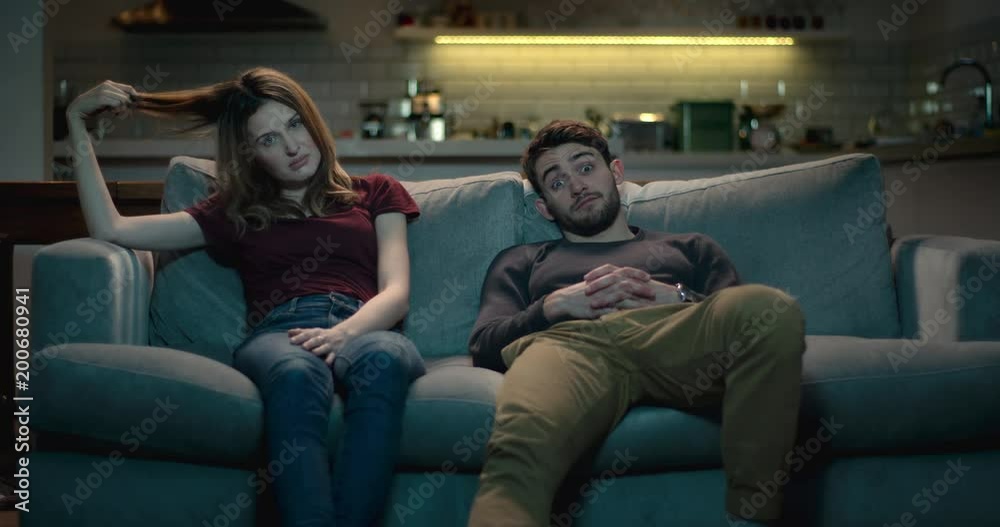 Extremely bored and frustrated couple sitting on a sofa at home struggle to watch a terrible movie..