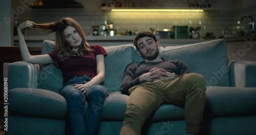 Extremely bored and frustrated couple sitting on a sofa at home struggle to watch a terrible movie..