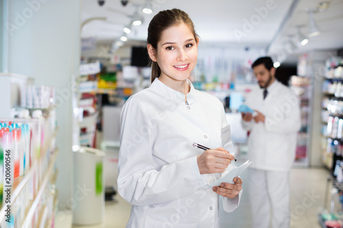 Female specialist is attentively stocktaking medicines with notebook near shelves in pharmacy.