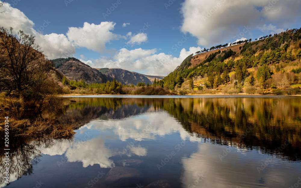 Irish Landscape - Blue sky with clouds and mountains reflecting in the calm water of the Lower Lake at the heart of the Glendalough Valley in Wicklow National Park in Ireland. 