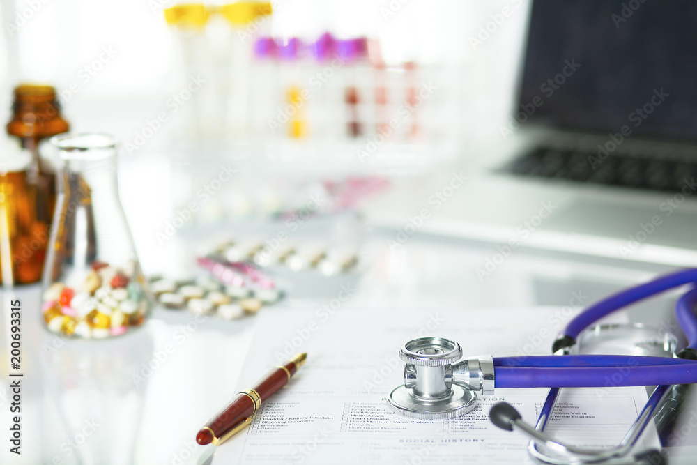 closeup of the desk of a doctors office with a stethoscope in the foreground and a bottle with pills in the background closeup of the desk of a doctors office with a stethoscope in the foreground and a bottle with pills in the background
