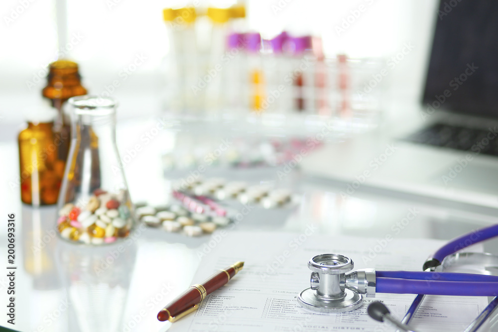 closeup of the desk of a doctors office with a stethoscope in the foreground and a bottle with pills in the background closeup of the desk of a doctors office with a stethoscope in the foreground and a bottle with pills in the background