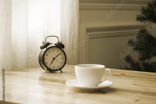 A cup of coffee and a pot of plants on a wooden table by the window. Focus on clock.