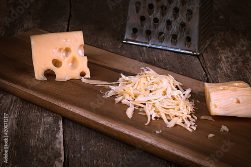 cheese on piece and grated and a used metal grater on a wooden cutting board against a dark slate background with copy space