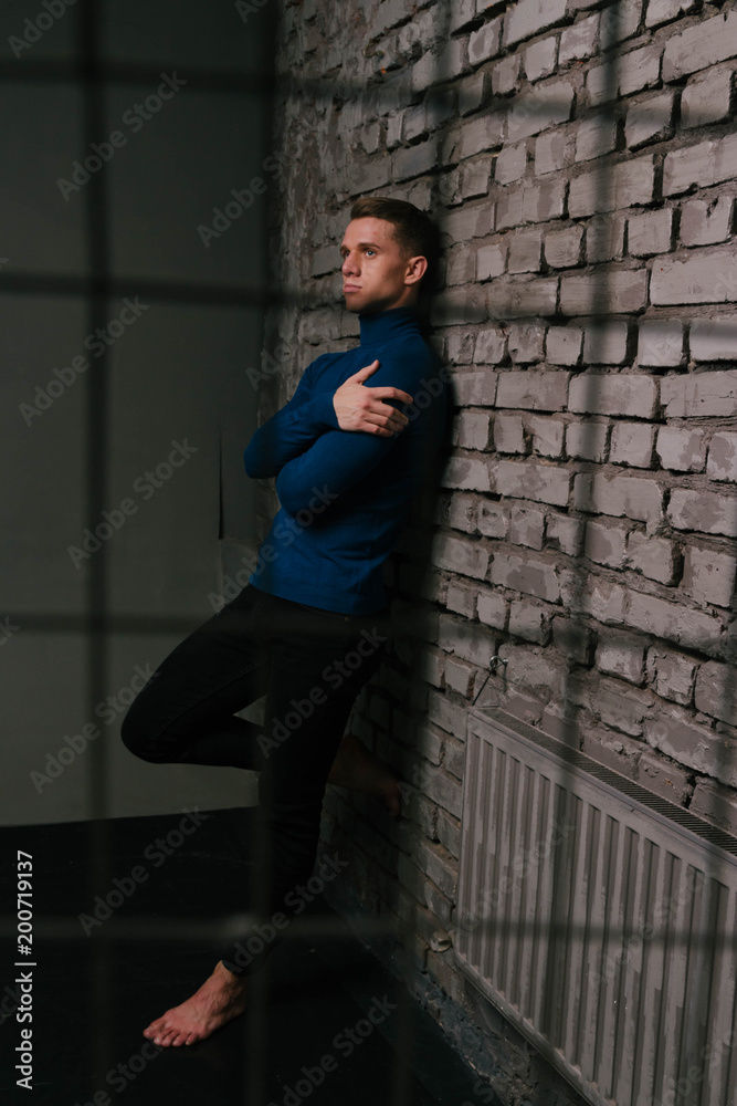 white attractive man in a blue sweater and black pants, barefoot stands  near a stone wall, behind bars, a prisoner Stock Photo | Adobe Stock