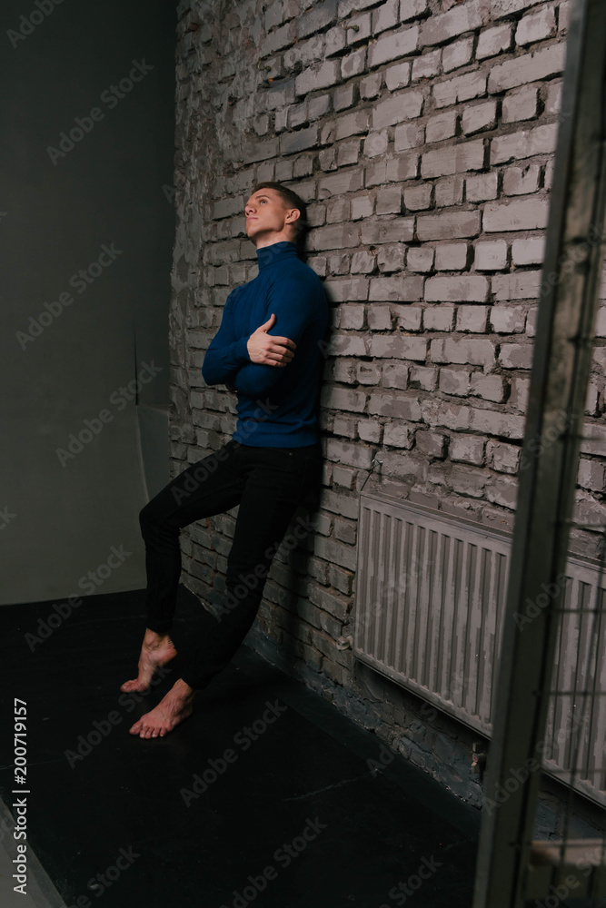 white attractive man in a blue sweater and black pants, barefoot stands near a stone wall, behind bars, a prisoner