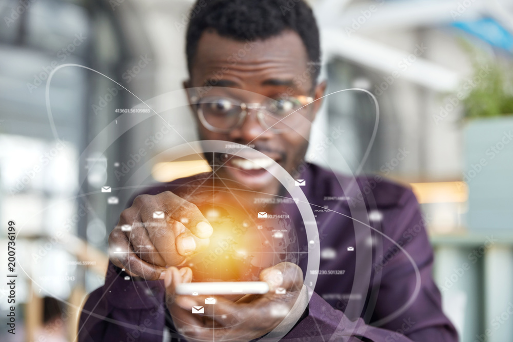African male enterpreneur holds modern cell phone with futuristic mobile application in hands, sends messages, has overjoyed expression. Double exposure and visual effects. Focus on electronic device
