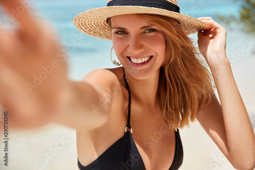 People, lifestyle, happiness and summer time concept. Lovely young smiling woman with cheerful expression poses for making selfie against azure sea background, happy to have good unforgettable rest
