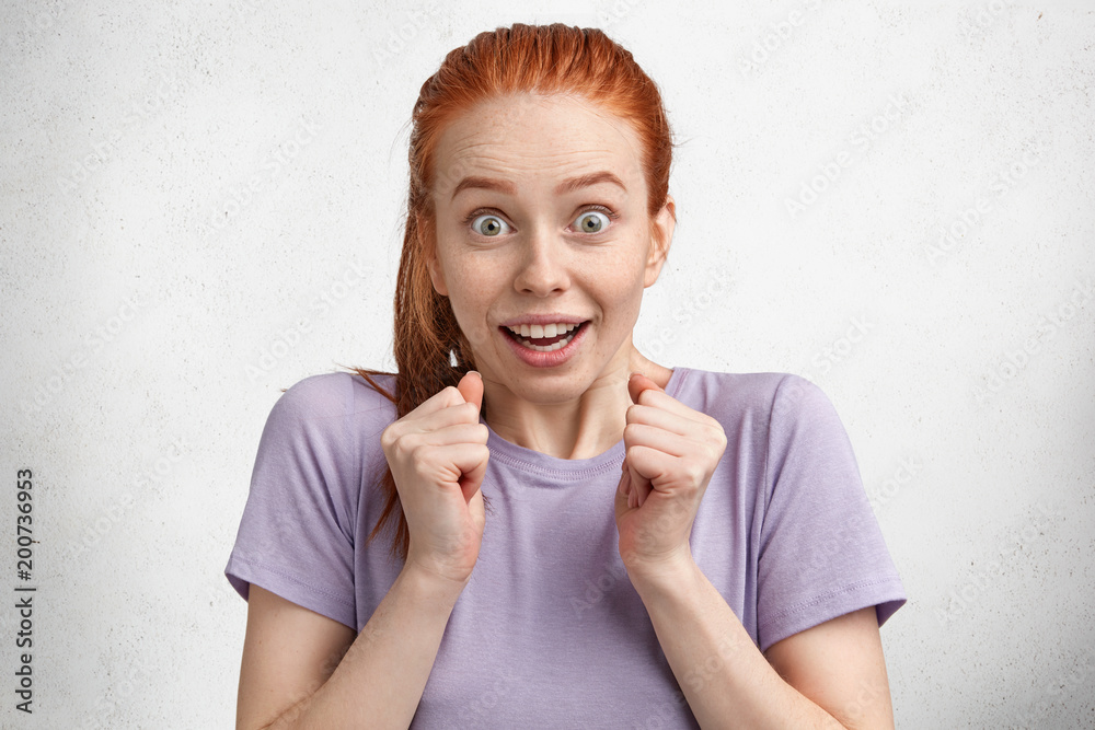 Overjoyed excited ginger young female model in purple casual t shirt, gestures actively, keeps hands in fists, demonstrates surprised reaction on what she hears, stares with bated breath at camera Overjoyed excited ginger young female model in purple casual t shirt, gestures actively, keeps hands in fists, demonstrates surprised reaction on what she hears, stares with bated breath at camera