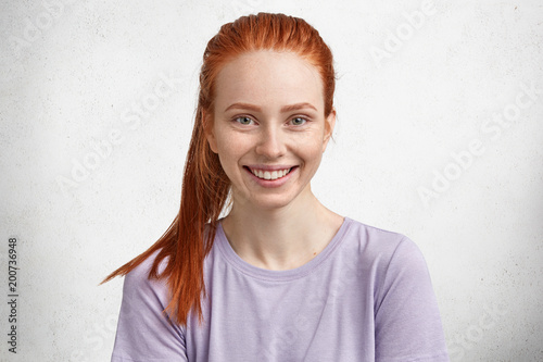 Red haired beautiful delighted female model being in high spirit after successful day, has freckled skin and ginger pony tail, poses against white concrete background. Skin care, beauty concept