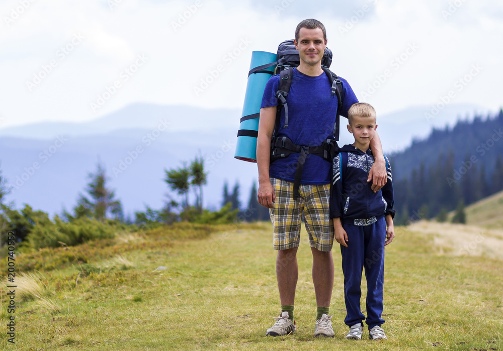 Father and son with backpacks hiking together in scenic summer green mountains. Dad and child standing enjoying landscape mountain view. Active lifestyle, family relations, weekend activity concept.