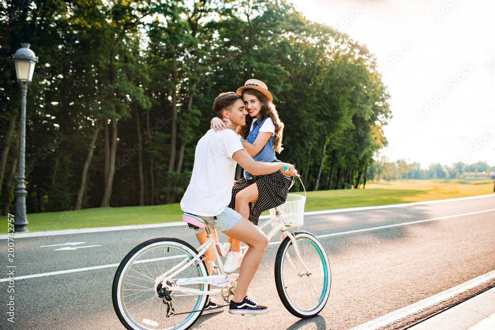 Full-length photo of romantic couple in love on bike on road on sunset. Handsome guy is driving a bike, girl with long curly hair in hat sitting on the handlebars, hugs him and smiles to camera. Full-length photo of romantic couple in love on bike on road on sunset. Handsome guy is driving a bike, girl with long curly hair in hat sitting on the handlebars, hugs him and smiles to camera.