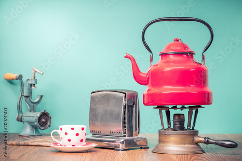 Retro classic red kettle on gas stove, a cup of tea, outdated bread toaster, kitchen board and vintage manual meat chopper on oak wooden table in front mint green background. Old style filtered photo