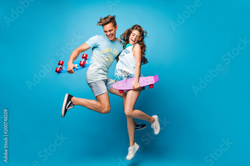 Young couple of handsome guy and pretty girl with long curly hair is jumping on blue background in studio. They wear T-shirts, jeans short, hold skateboards in hands.
