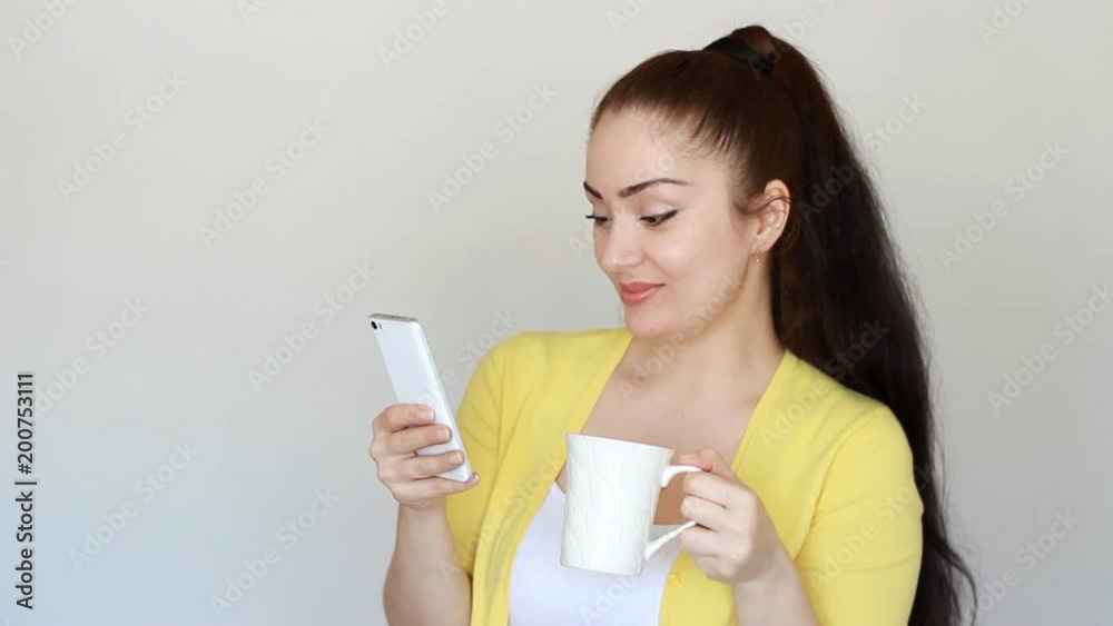 Portrait of a beautiful brunette girl who drinks a hot drink from a cup and holds a mobile phone in his hand. Young woman reads social network and looks internet on smartphone screen and smiles. Portrait of a beautiful brunette girl who drinks a hot drink from a cup and holds a mobile phone in his hand. Young woman reads social network and looks internet on smartphone screen and smiles.