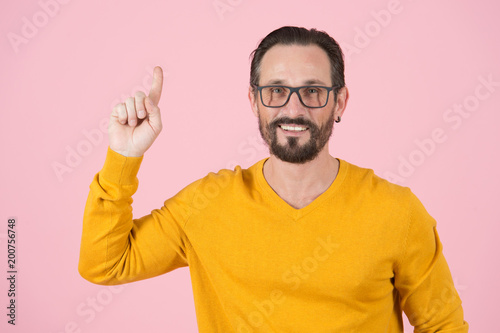 Finger up by bearded smiling man in glasses pointing up on pink background. Happy man with finger up. Wise Man in glasses notices up with finger. Clever guy in orange on pink background. © ReyRomMedia