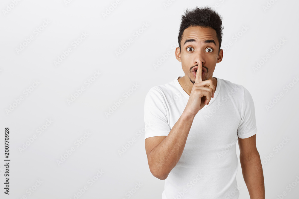 Shh, this is secret. Concerned good-looking african male model with afro haircut in casual white t-shirt, showing shush gesture with index finger over mouth, worrying, asking to keep voice down Shh, this is secret. Concerned good-looking african male model with afro haircut in casual white t-shirt, showing shush gesture with index finger over mouth, worrying, asking to keep voice down