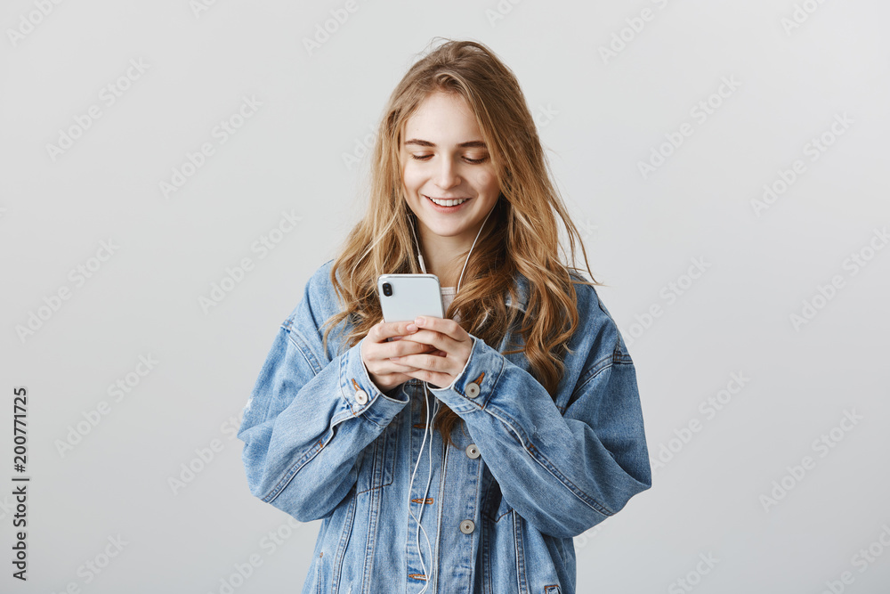 Watching video on social network. Indoor shot of good-looking european female with blond hair, wearing denim jacket, typing message in smartphone, smiling curiously and listening music in earphones Watching video on social network. Indoor shot of good-looking european female with blond hair, wearing denim jacket, typing message in smartphone, smiling curiously and listening music in earphones