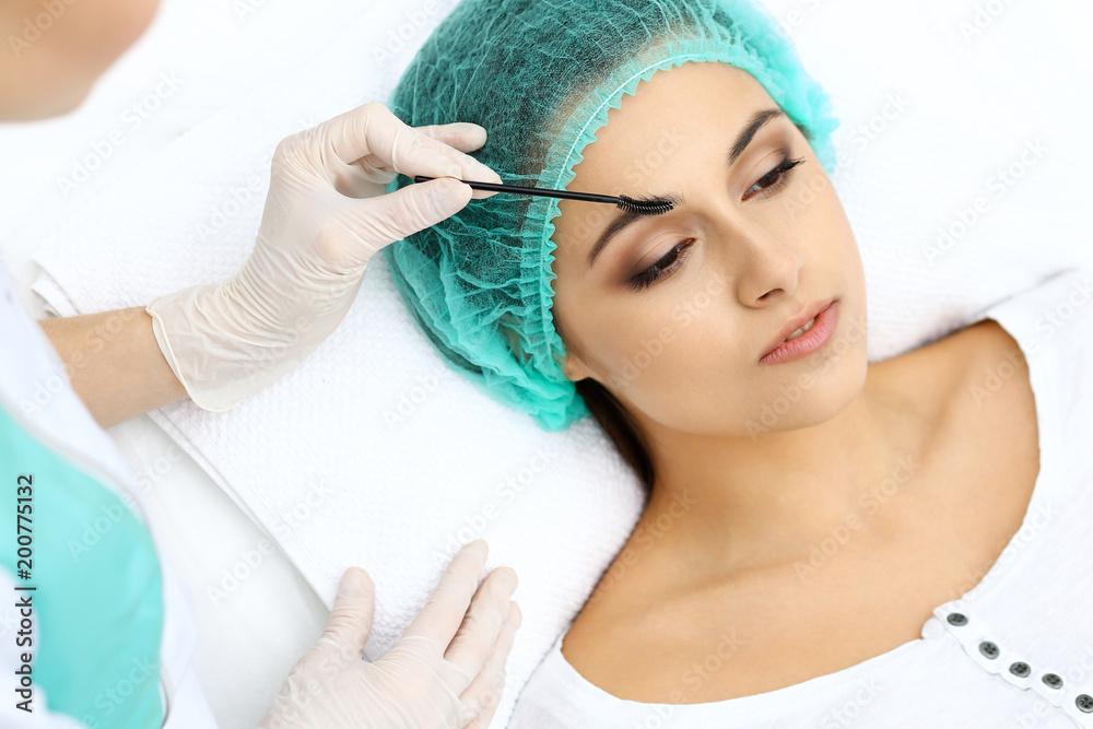 Professional beautician is doing cosmetic procedure at light medical background touching patient's face with brush, closeup. Cosmetology treatment Professional beautician is doing cosmetic procedure at light medical background touching patient's face with brush, closeup. Cosmetology treatment