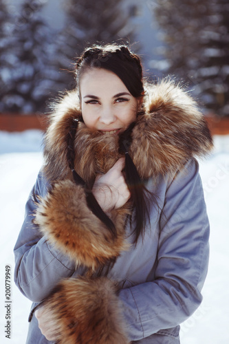 Beautiful brunette girl with two pigtails in a blue coat with a brown fluffy fur collar in the winter on nature. Snowy spruce on the background. photo