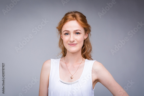 Closeup portrait young, beautiful business woman, student with lred, curly hair and freckles on face on gray background in the studio. Dressed in white blouse with short sleeves about open shoulders