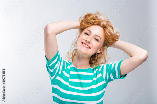 Close-up portrait of a young, beautiful woman with red curly hair in a summer dress with strips of blue in the studio on a gray background. Theme of summer vacation, tourism and summer clothes
