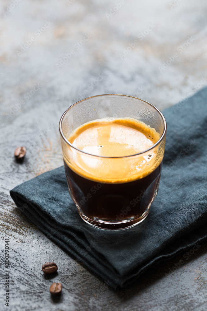 Glass cup of fresh coffee with thick golden foam froth on dark rustic background, espresso americano on black tissue with spoon of brown sugar and coffee beans, vertical copy space Glass cup of fresh coffee with thick golden foam froth on dark rustic background, espresso americano on black tissue with spoon of brown sugar and coffee beans, vertical copy space