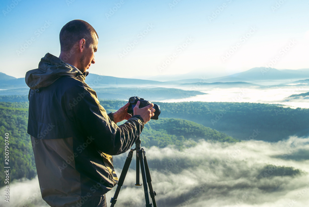 Photographer takes photos with camera on tripod on rocky mountain peak. Beautiful misty sunrise and valley view over clouds.