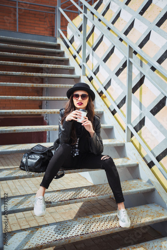Young stylish woman in a city street sitting and drinking coffee or tea . © Elena Kratovich