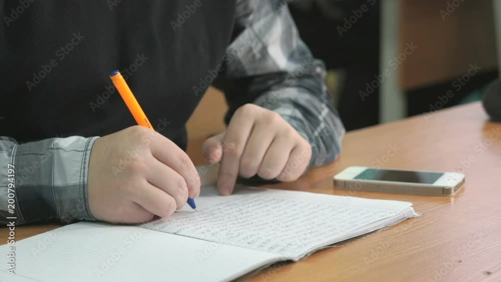Unknown man in grey shirt with black pullover sitting at desk at lesson at university writes text using ball pen in notebook. Student looks at silver mobile phone for important information Unknown man in grey shirt with black pullover sitting at desk at lesson at university writes text using ball pen in notebook. Student looks at silver mobile phone for important information